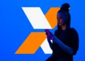 Mulher usa celular diante de marca em formato de X laranja e branco que representa o programa Desenrola.