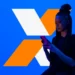 Mulher usa celular diante de marca em formato de X laranja e branco que representa o programa Desenrola.