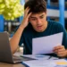 Estudante preocupado consulta documento ao lado de notebook, representando o prazo final das vagas remanescentes do Fies 2026