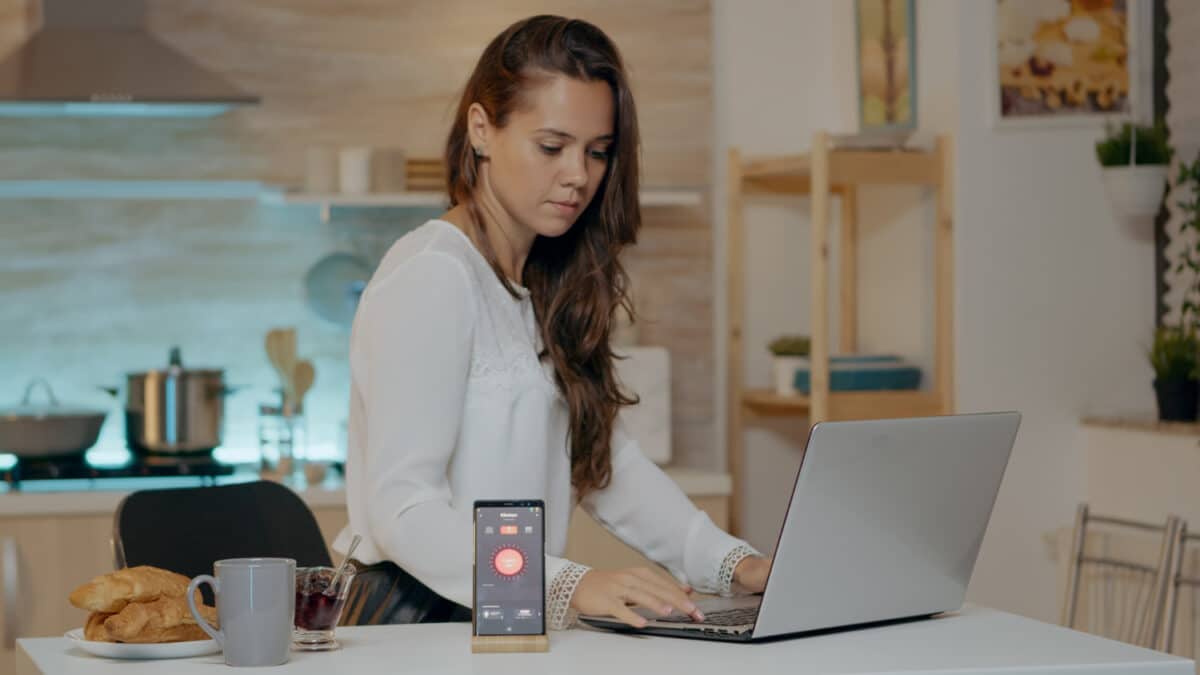 Mulher estuda em casa pelo notebook na cozinha, ao lado de celular e xícara de café