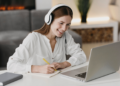 Mulher ruiva sorridente com fone branco faz anotações em caderno enquanto olha para laptop em ambiente doméstico.