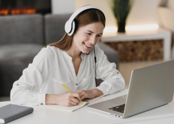 Mulher ruiva sorridente com fone branco faz anotações em caderno enquanto olha para laptop em ambiente doméstico.