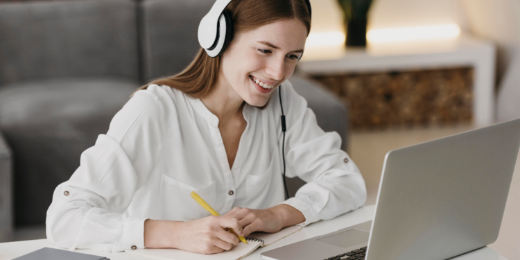 Mulher ruiva sorridente com fone branco faz anotações em caderno enquanto olha para laptop em ambiente doméstico.