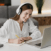 Mulher ruiva sorridente com fone branco faz anotações em caderno enquanto olha para laptop em ambiente doméstico.