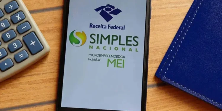 Tela de celular exibe logos da Receita Federal e do Simples Nacional para Microempreendedor Individual ao lado de calculadora cinza sobre mesa de madeira.