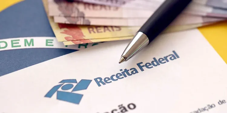Folha com timbre da Receita Federal repousa sobre superfície amarela ao lado de notas de real e caneta esferográfica preta apoiada no documento