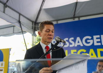 Homem de terno preto e gravata vermelha discursa ao microfone segurando documentos em evento oficial
