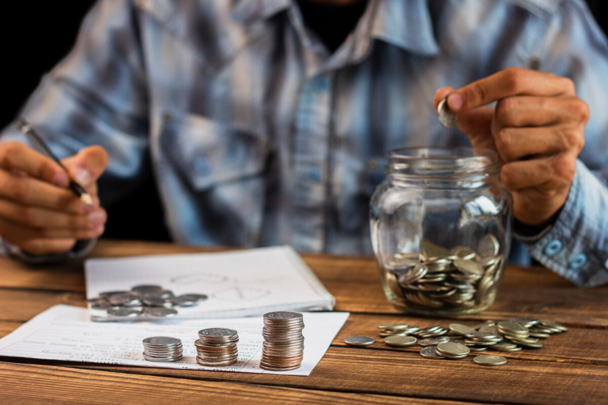 Homem de camisa xadrez azul guardando moedas em pote de vidro sobre mesa de madeira com documentos e pilhas de moedas organizadas