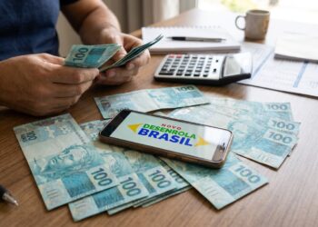 Homem conta cédulas de cem reais ao lado de celular que exibe a marca do Novo Desenrola Brasil e uma calculadora.