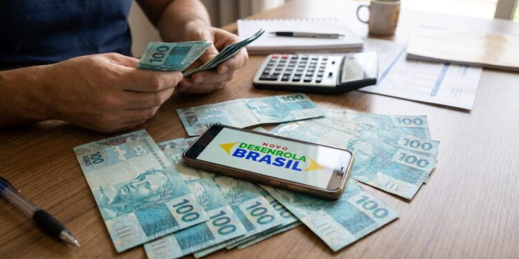 Homem conta cédulas de cem reais ao lado de celular que exibe a marca do Novo Desenrola Brasil e uma calculadora.
