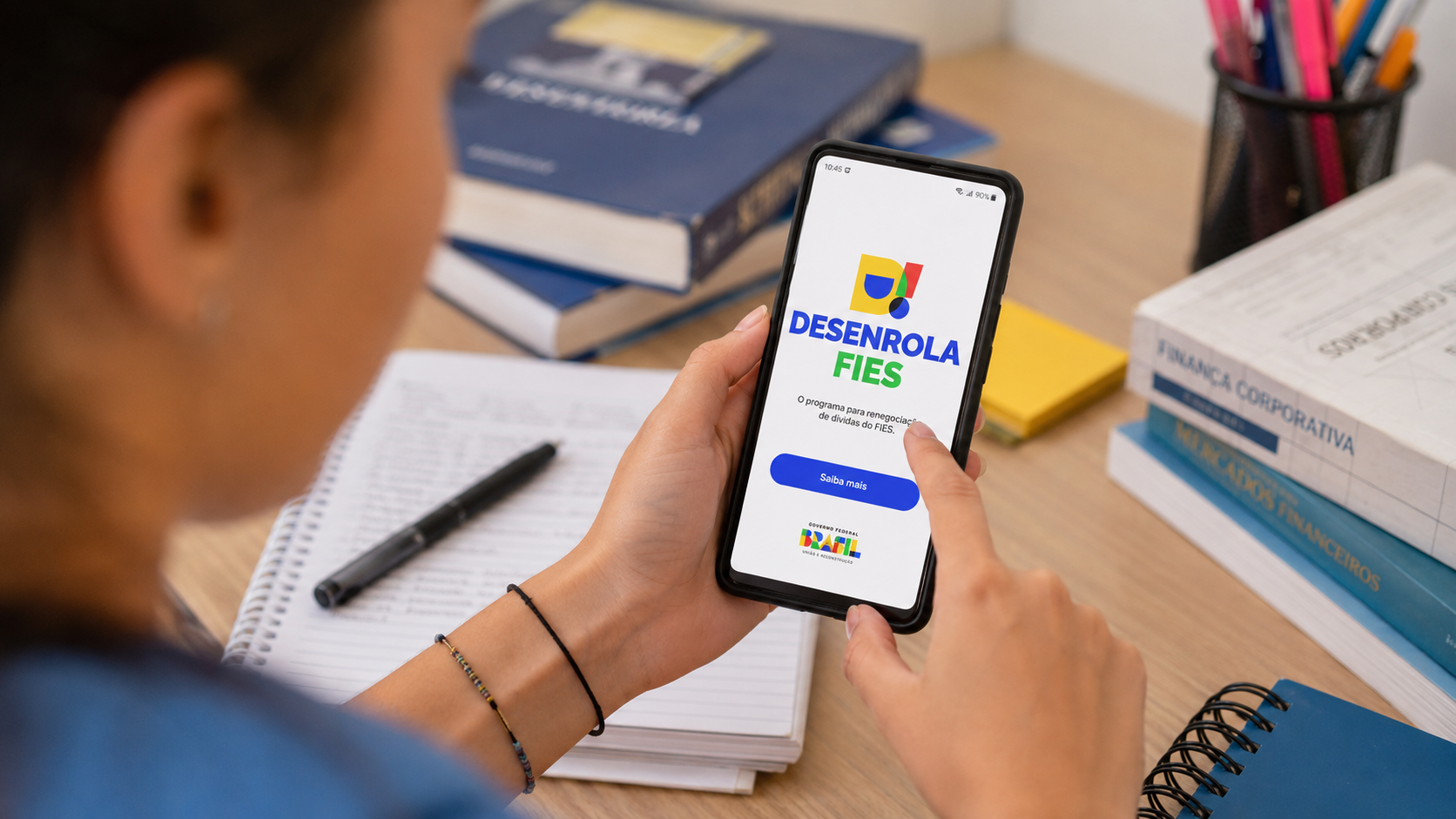 Jovem segura smartphone exibindo tela do Desenrola FIES com botão azul "Saiba mais" e logo do Governo Federal