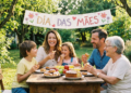 Família com três gerações sorri em volta de mesa rústica com bolos, frutas e suco, sob faixa escrita Dia das Mães.