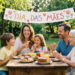 Família com três gerações sorri em volta de mesa rústica com bolos, frutas e suco, sob faixa escrita Dia das Mães.