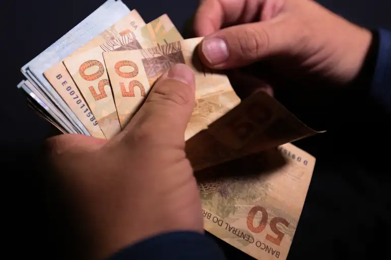 Mãos contando cédulas de cinquenta reais brasileiros em close-up sobre fundo escuro
