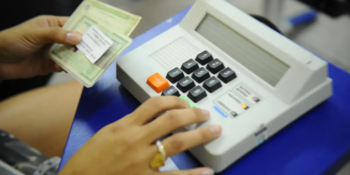 Eleitor segurando título de eleitor diante de urna eletrônica durante o processo de votação no Brasil, representando o direito ao voto.