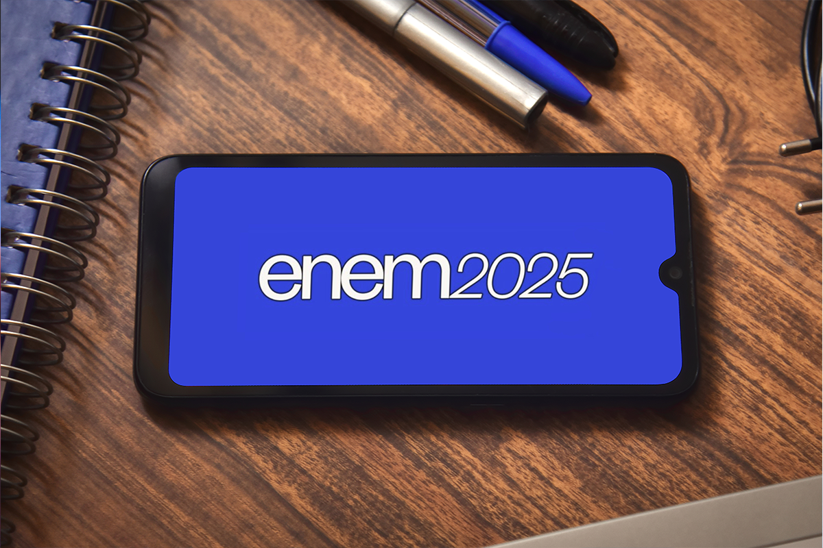 Smartphone com tela azul exibindo "enem2025" sobre mesa de madeira com cadernos espirais e canetas.
