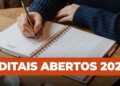 Mãos escrevendo em agenda 2026 com caneta prata sobre mesa de madeira, destacando texto editais abertos 2026