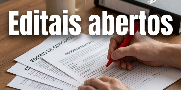 Mão segurando caneta vermelha preenche formulários de editais de concurso e processo seletivo sobre mesa de madeira.