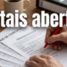 Mão segurando caneta vermelha preenche formulários de editais de concurso e processo seletivo sobre mesa de madeira.