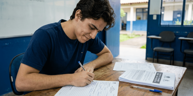 : Estudante preenche ficha de inscrição em sala de aula com cadernos e materiais sobre a mesa