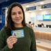 Mulher de blusa verde sorri segurando a nova Carteira de Identidade Nacional diante dos guichês de atendimento em centro comercial