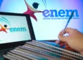 Mão segurando caneta sobre cadernos espirais, laptop exibindo logo do ENEM em ambiente de estudo azulado.