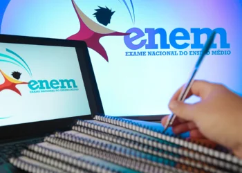 Mão segurando caneta sobre cadernos espirais, laptop exibindo logo do ENEM em ambiente de estudo azulado.