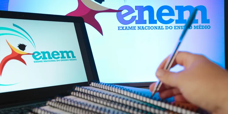 Mão segurando caneta sobre cadernos espirais, laptop exibindo logo do ENEM em ambiente de estudo azulado.