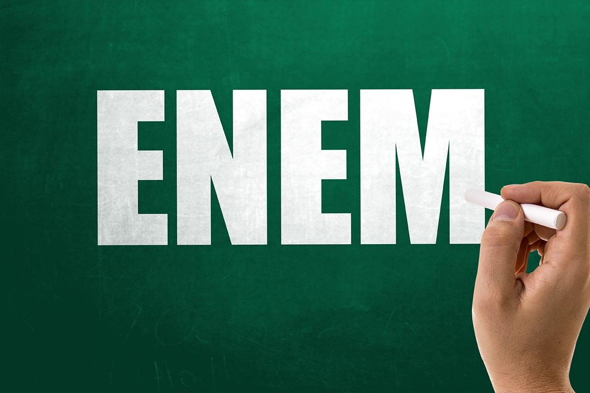 Palavra “Enem” em quadro verde, com mão segurando giz, representa a expectativa pela abertura das inscrições do exame em 2026.