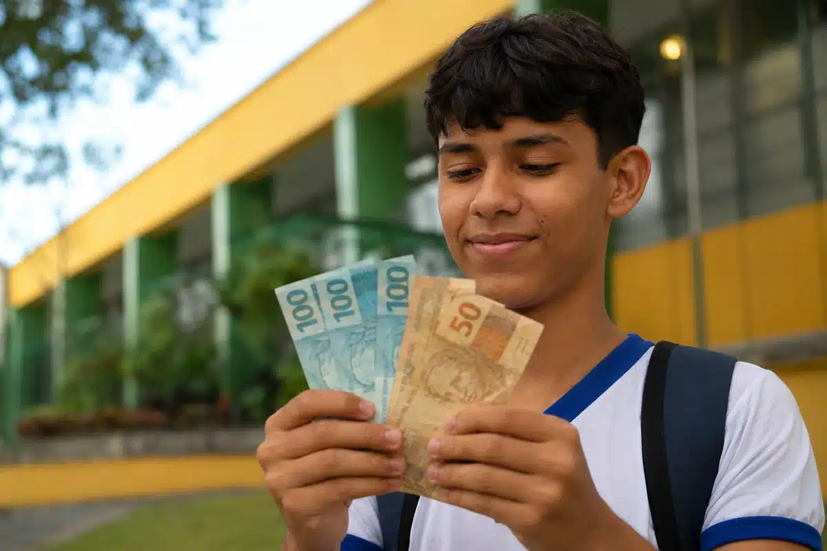 Estudante da rede pública segura cédulas de dinheiro em frente à escola, em referência ao bônus de R$ 200 do Enem 2026.