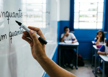 Professor escreve as palavras enquanto e em quanto em quadro branco enquanto estudantes uniformizados acompanham a aula ao fundo