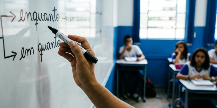 Professor escreve as palavras enquanto e em quanto em quadro branco enquanto estudantes uniformizados acompanham a aula ao fundo