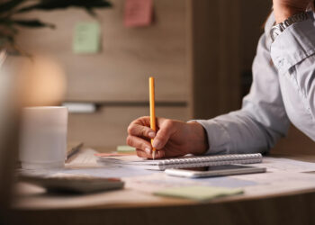 Pessoa de camisa social escreve a lápis em caderno espiral sobre mesa de escritório com caneca branca e papéis ao redor