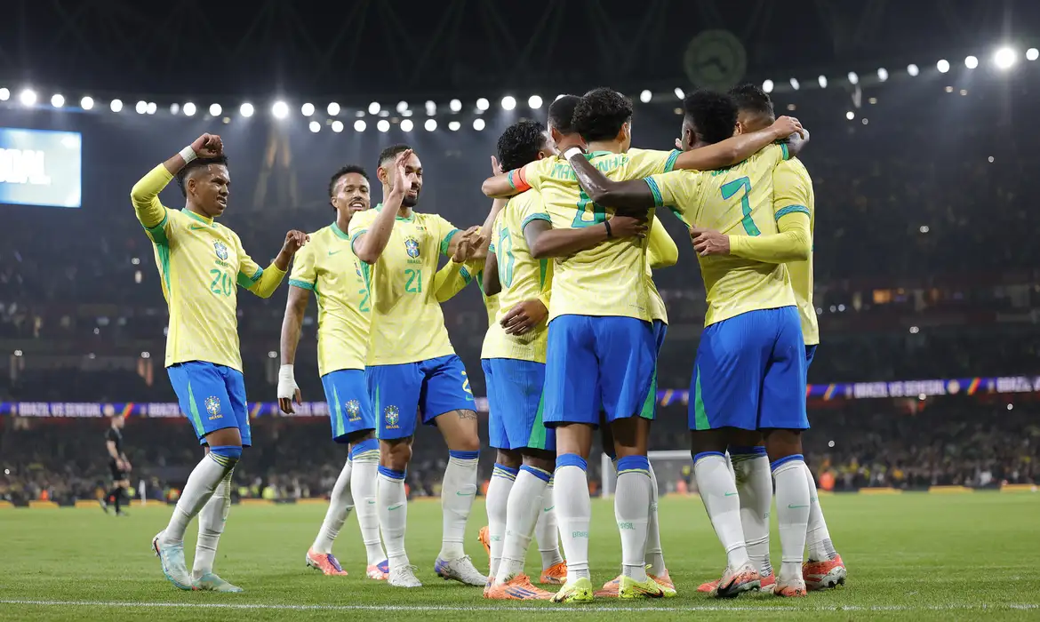 Jogadores da Seleção Brasileira reunidos em abraço coletivo no campo durante comemoração de gol em partida noturna em estádio