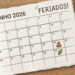 Calendário de junho 2026 sobre mesa de madeira com fogueira no dia 24 e milho no dia 29, xícara de café e caneta.