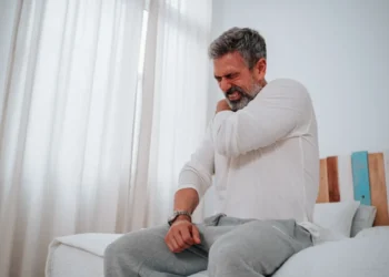 Homem sentado na cama com expressão de dor no ombro, ilustrando sintomas de fibromialgia relacionados ao reconhecimento para auxílio-doença e aposentadoria.