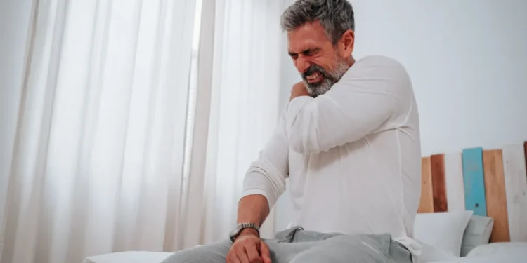 Homem sentado na cama com expressão de dor no ombro, ilustrando sintomas de fibromialgia relacionados ao reconhecimento para auxílio-doença e aposentadoria.