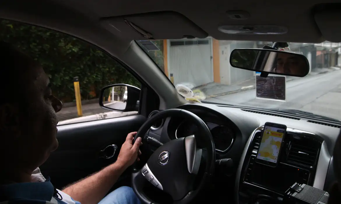 Motorista ao volante de um veículo em via urbana, com smartphone fixado no painel exibindo aplicativo de navegação, em referência ao novo sistema do Ministério dos Transportes para conectar alunos a instrutores de direção.