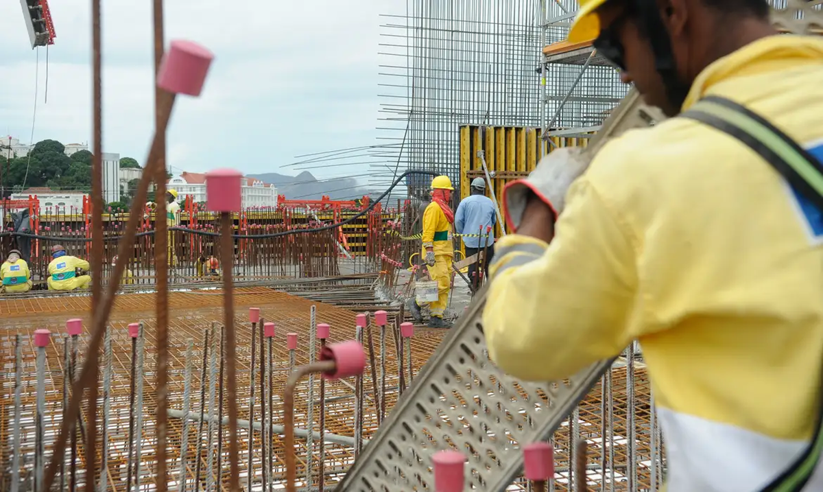 Operários da construção civil trabalhando em canteiro de obras, com destaque para trabalhador de capacete amarelo em primeiro plano