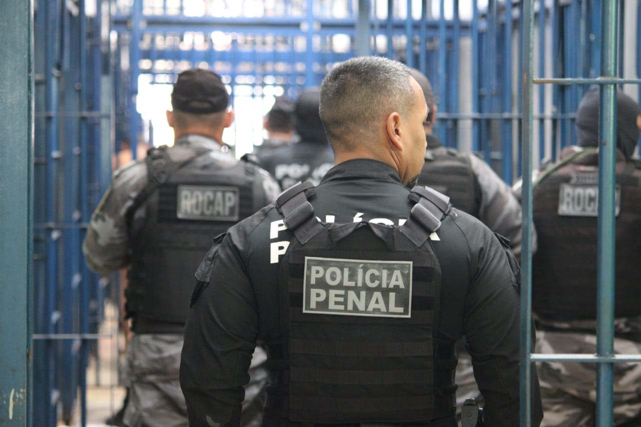 Policiais penais com coletes pretos identificados entram em corredor de unidade prisional com grades azuis