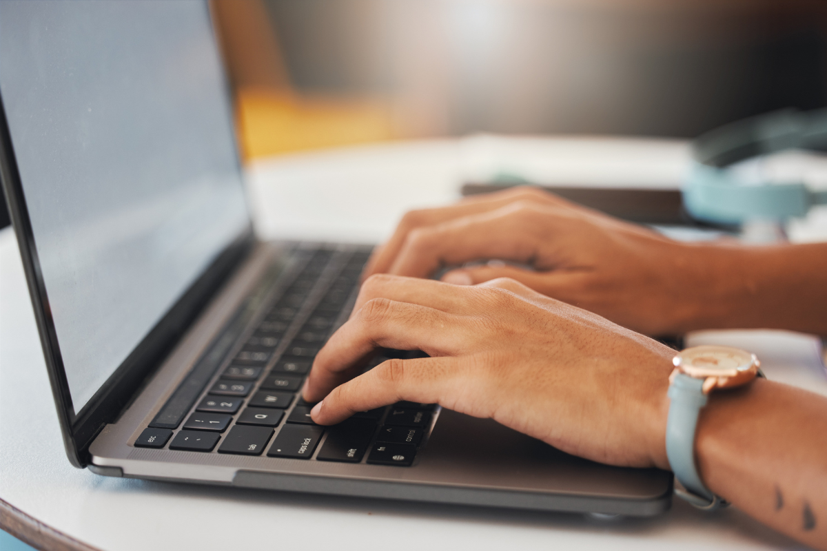 Mãos femininas digitando em teclado de MacBook cinza-espacial, usando relógio rosé com pulseira azul turquesa.