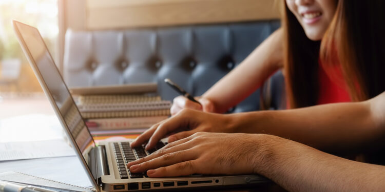 Estudante de óculos sorri ao lado de pessoa que digita em notebook durante inscrição no Enem 2026.