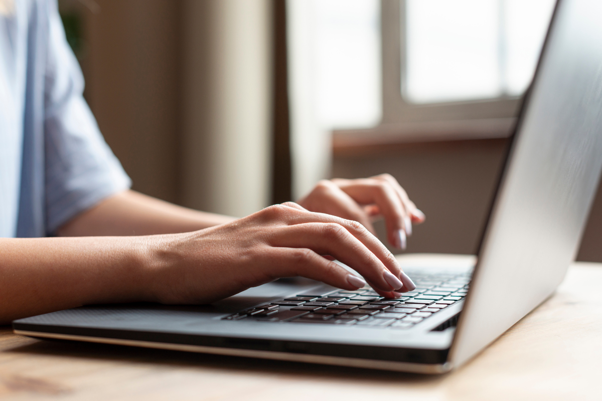 Mãos femininas com camisa branca digitam em teclado de notebook prateado sobre mesa de madeira clara.