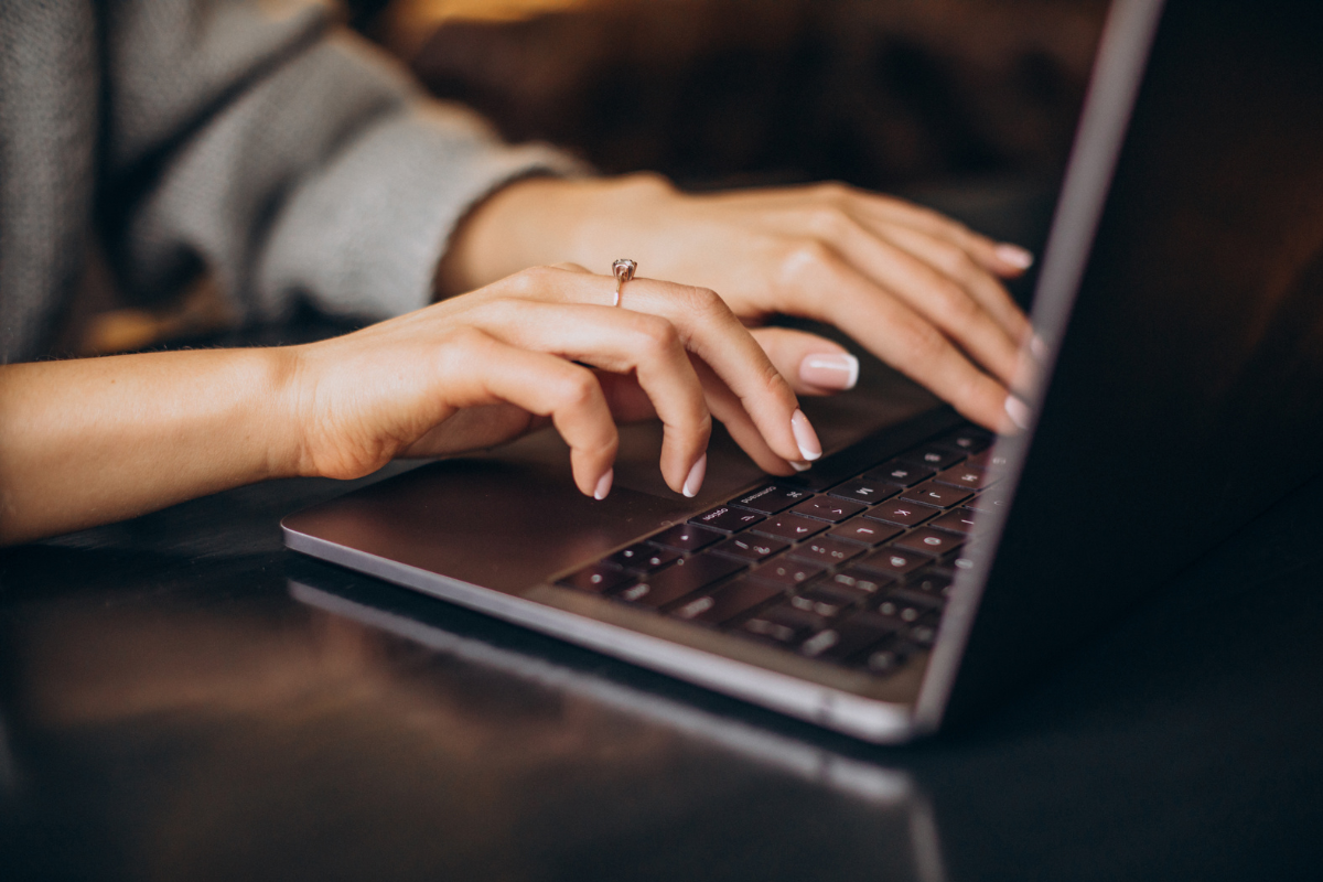 Mãos de mulher com unhas claras e anel digitando em teclado de MacBook prateado sobre mesa escura