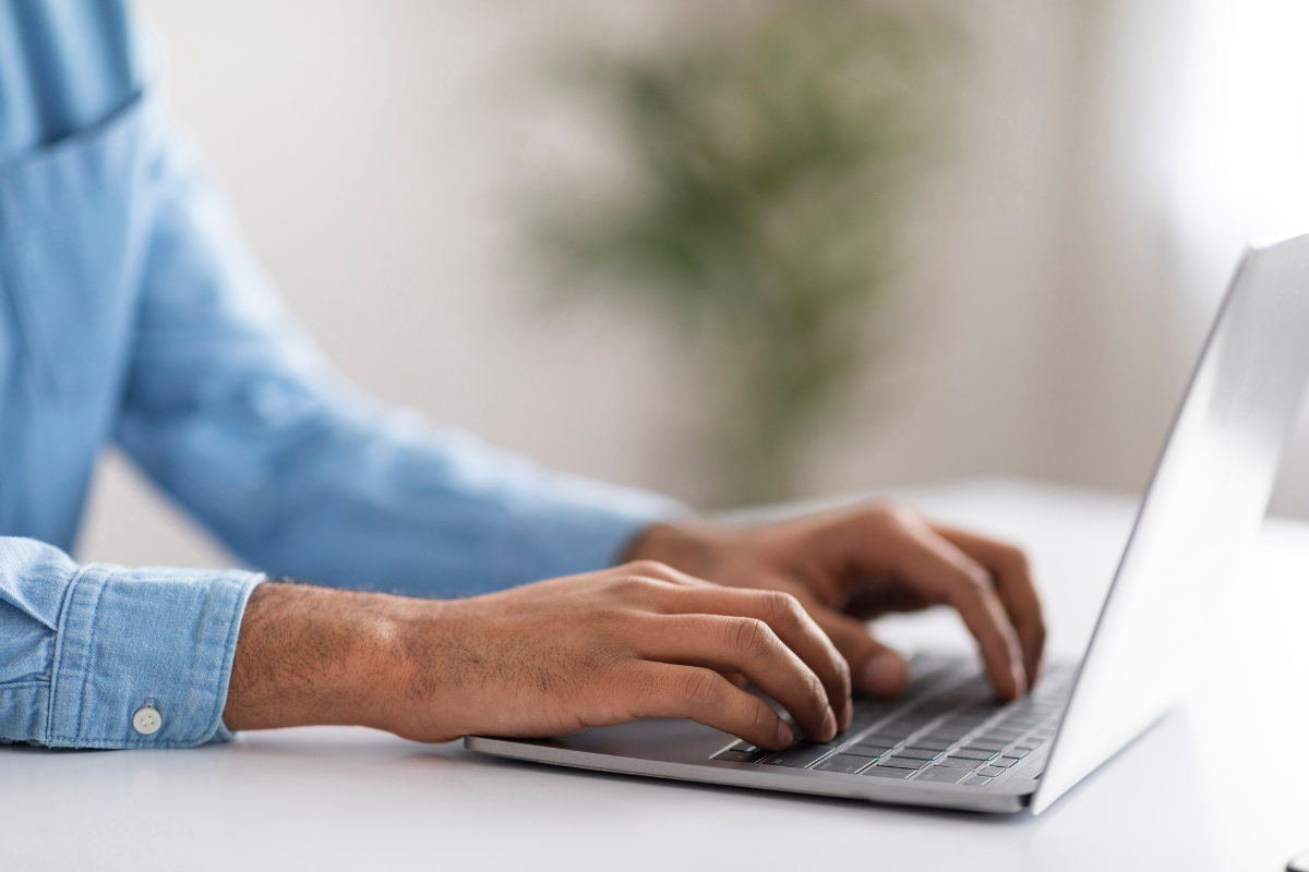 Mãos de homem com camisa jeans azul digitando em notebook cinza sobre mesa branca, planta desfocada ao fundo.