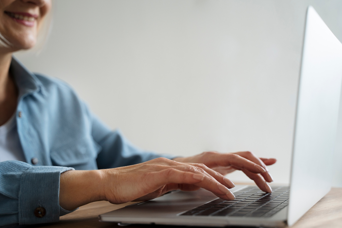 Mãos femininas digitando em laptop prateado sobre mesa de madeira, mulher de camisa azul sorrindo