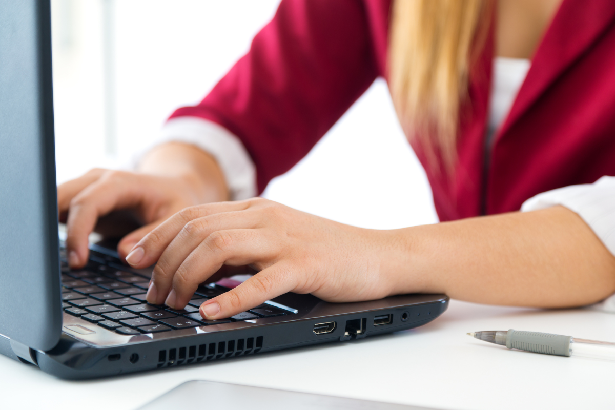Mãos femininas digitando em teclado de notebook preto, blazer vermelho e cabelo loiro visíveis