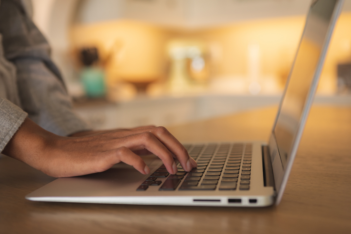 Mão feminina digitando em teclado de notebook prateado sobre mesa de madeira com iluminação quente ao fundo.