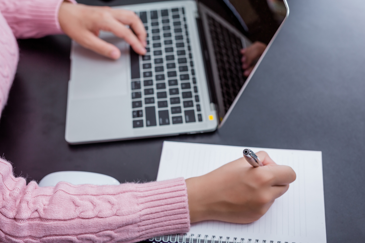 Mãos femininas com suéter rosa tricotado escrevendo em caderno pautado ao lado de laptop prateado sobre mesa preta.