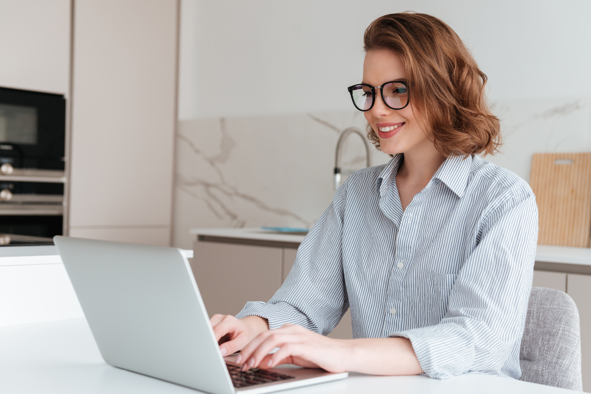 Mulher ruiva de óculos e camisa listrada digita sorrindo em laptop prateado na bancada de cozinha clara.